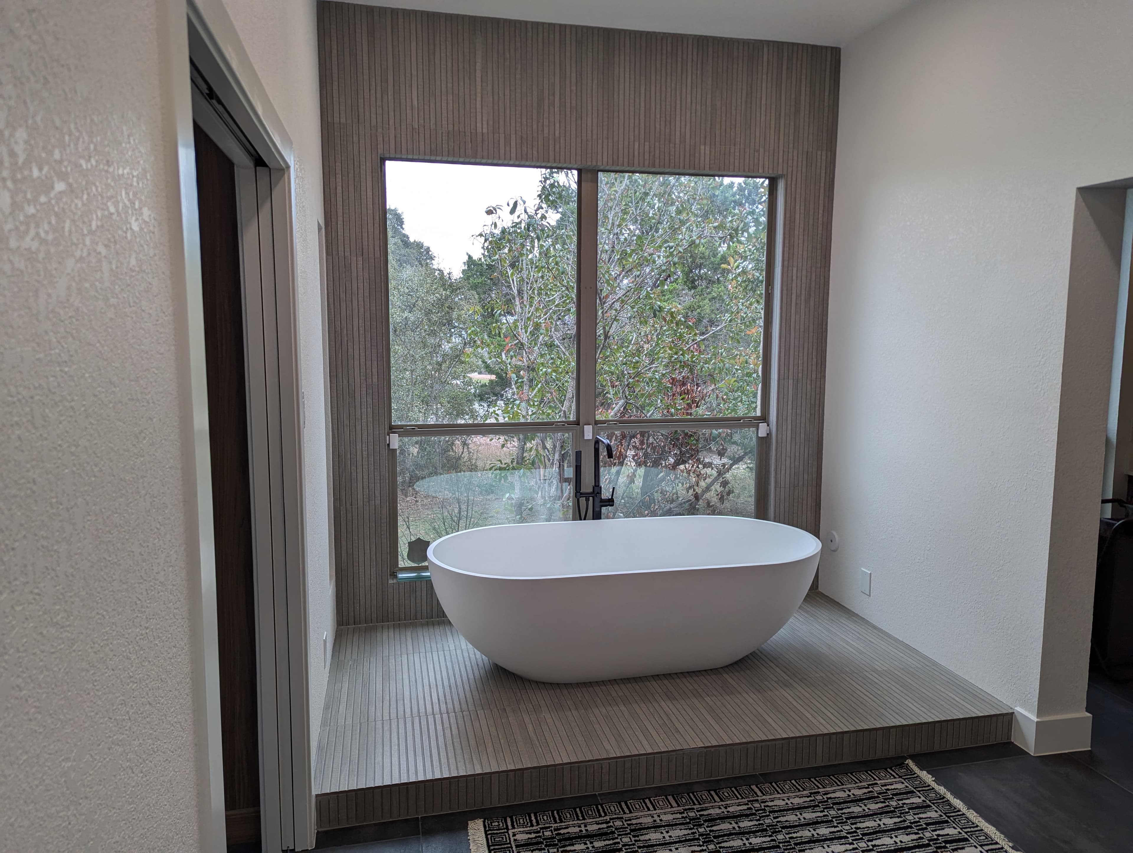Luxurious master bathroom centerpiece featuring a modern freestanding soaking tub positioned on a raised platform with gray tile detailing. The stunning textured fluted tile accent wall creates architectural interest, complemented by matte black floor-mount tub filler and large picture window with natural views.