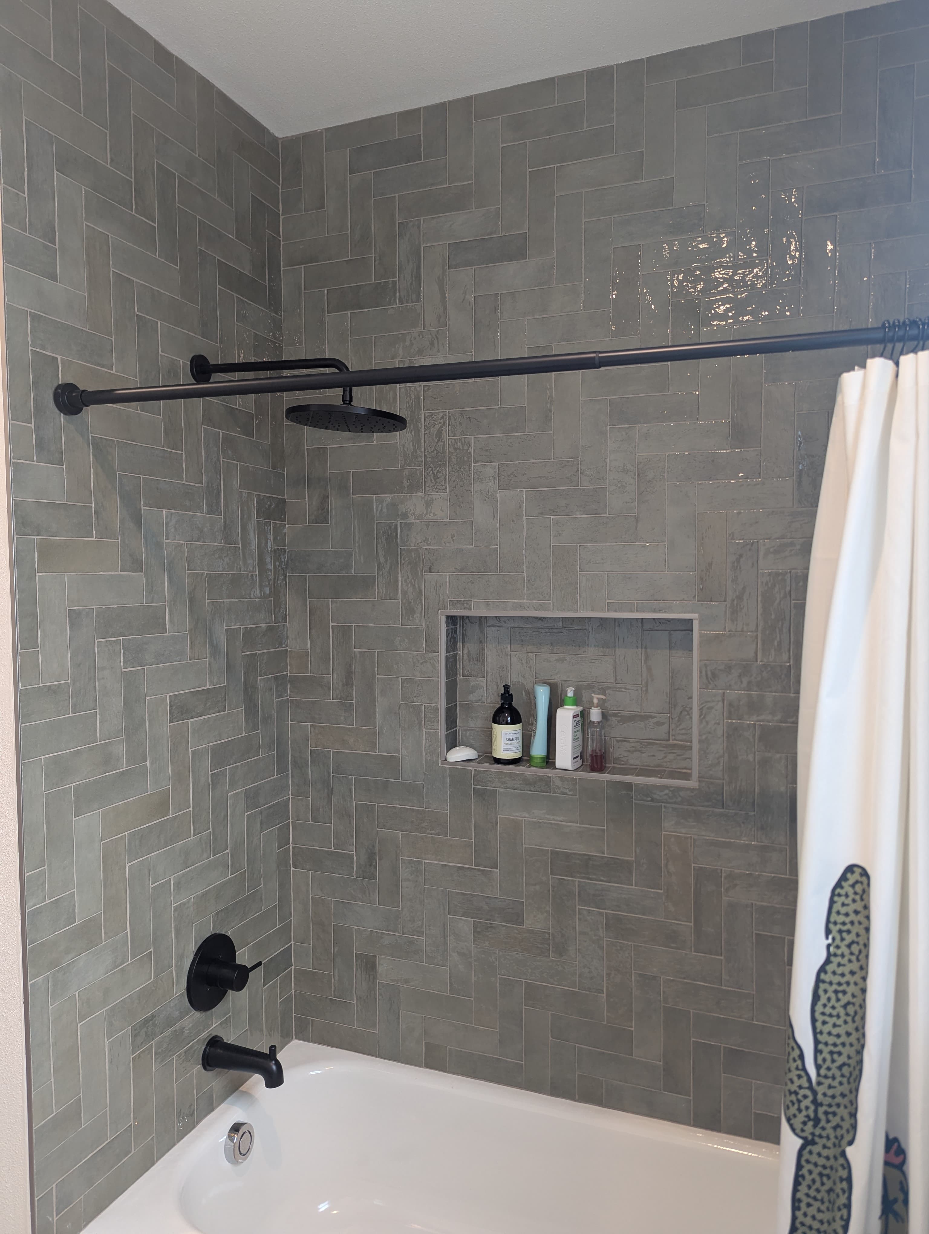 Modern tub-shower combo featuring sophisticated gray wood-look tile in a stylish herringbone pattern. The design includes a recessed shower niche for storage, matte black shower fixtures with rainfall showerhead, and sleek white shower curtain for a clean, contemporary aesthetic.