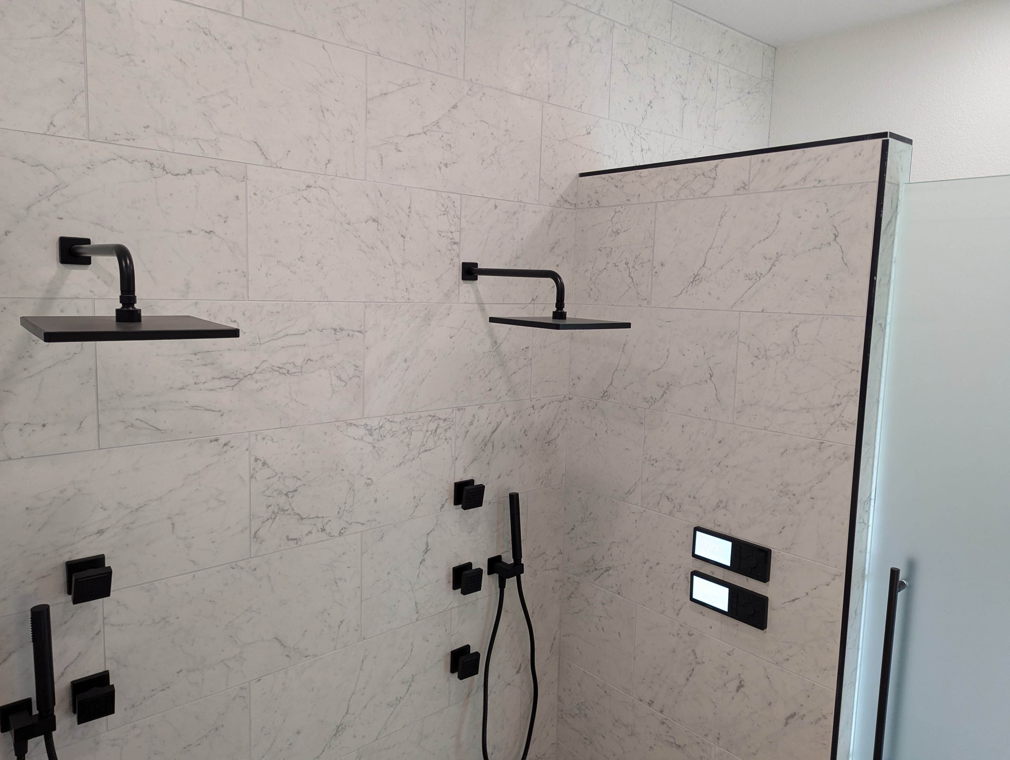 High-end walk-in shower featuring elegant large-format marble-look porcelain tile with natural veining throughout. This luxurious design includes dual rainfall showerheads, matte black fixtures, handheld shower wand, built-in body sprays, and frameless glass enclosure. A perfect example of modern luxury bathroom design.