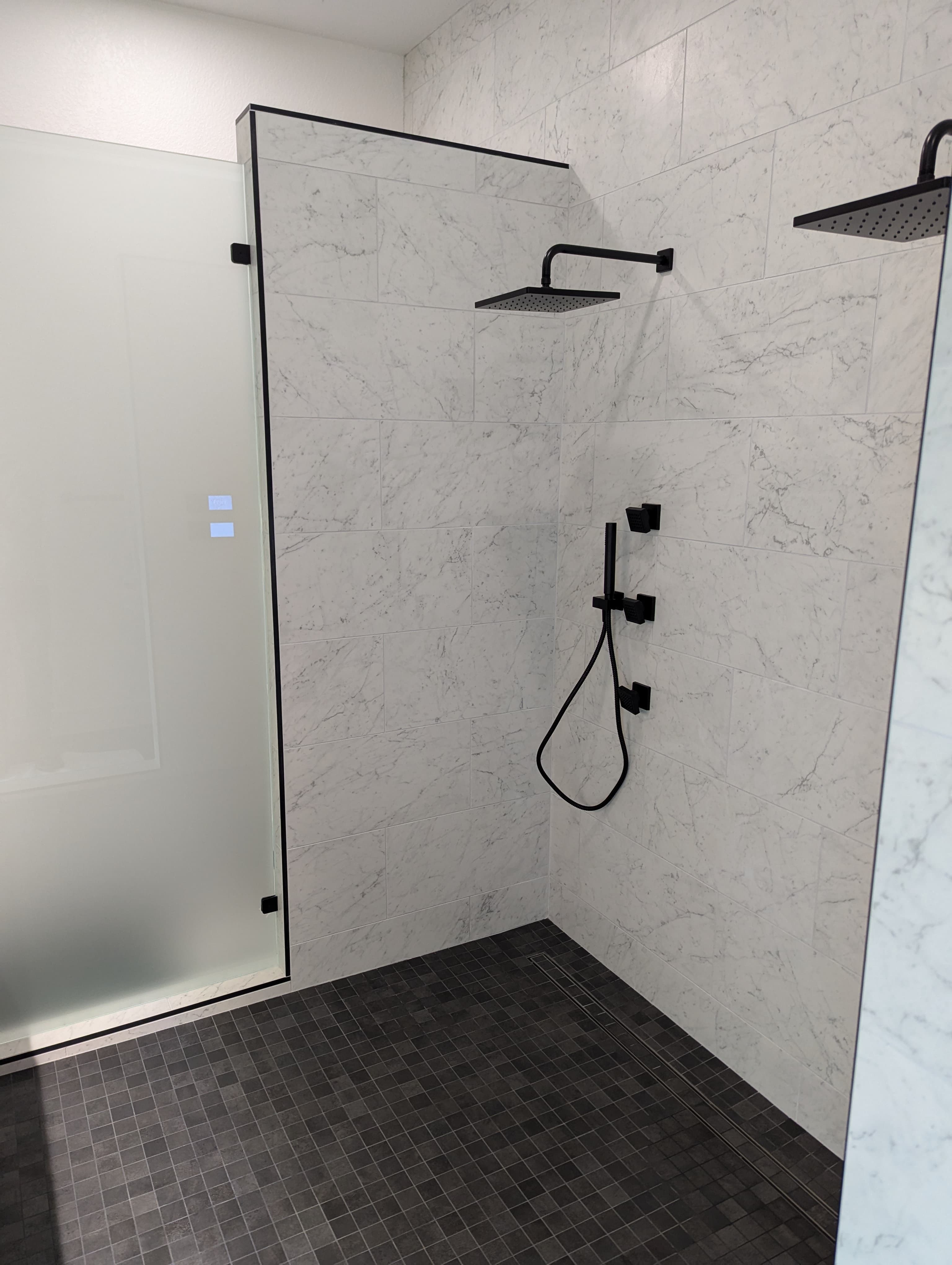 Contemporary walk-in shower with floor-to-ceiling marble-look tile, frameless glass door with matte black hardware, dual rainfall showerheads, and handheld shower attachment. The cohesive design showcases premium materials and modern fixtures, creating an elegant and functional bathing space.