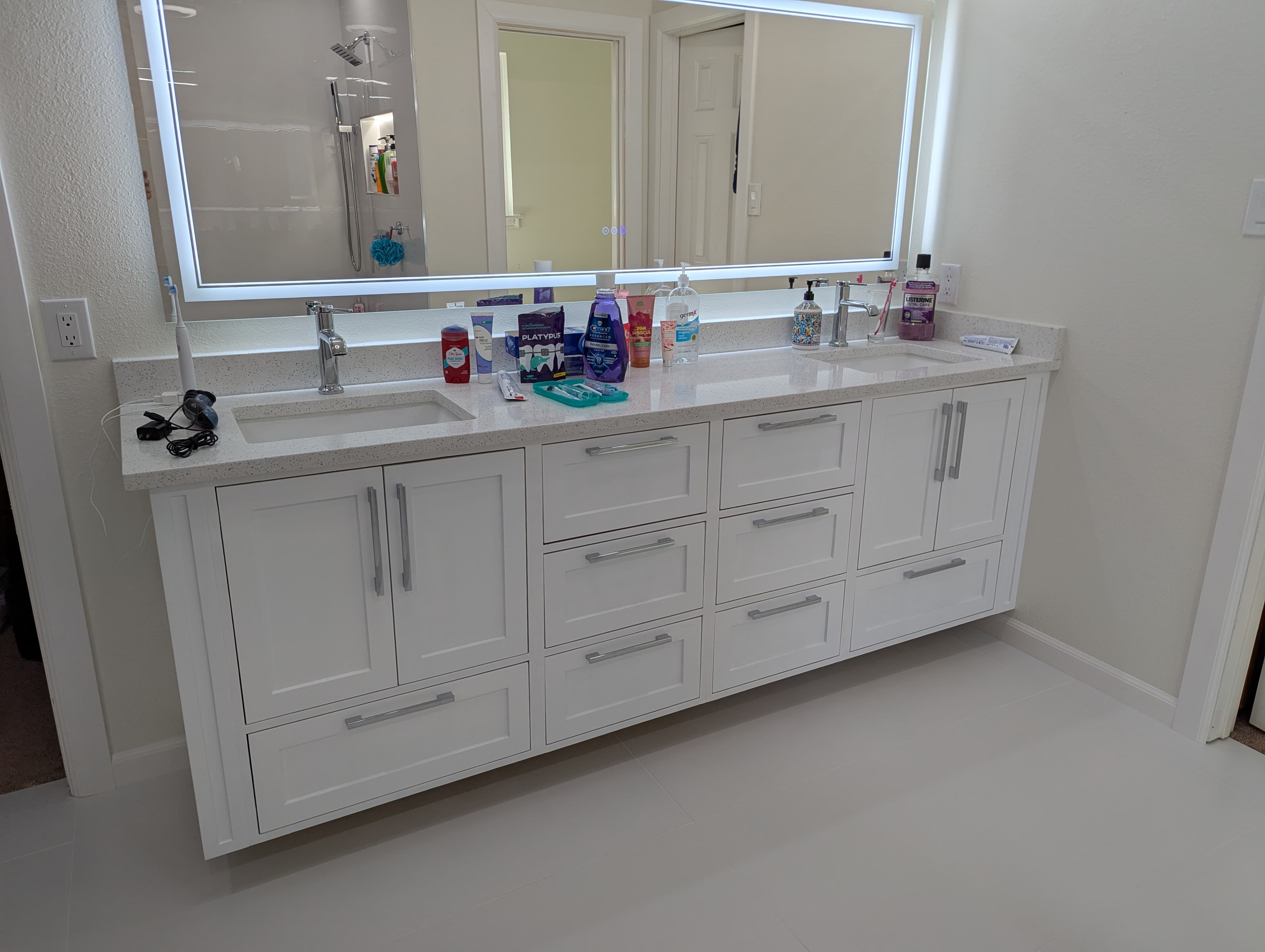 Stunning modern bathroom featuring a custom white double vanity with inset cabinet doors, premium quartz countertops, dual undermount sinks, and a full-width LED-backlit mirror. The clean white cabinetry includes multiple drawers and cabinet doors for optimal storage, complemented by contemporary chrome fixtures and large-format tile flooring.