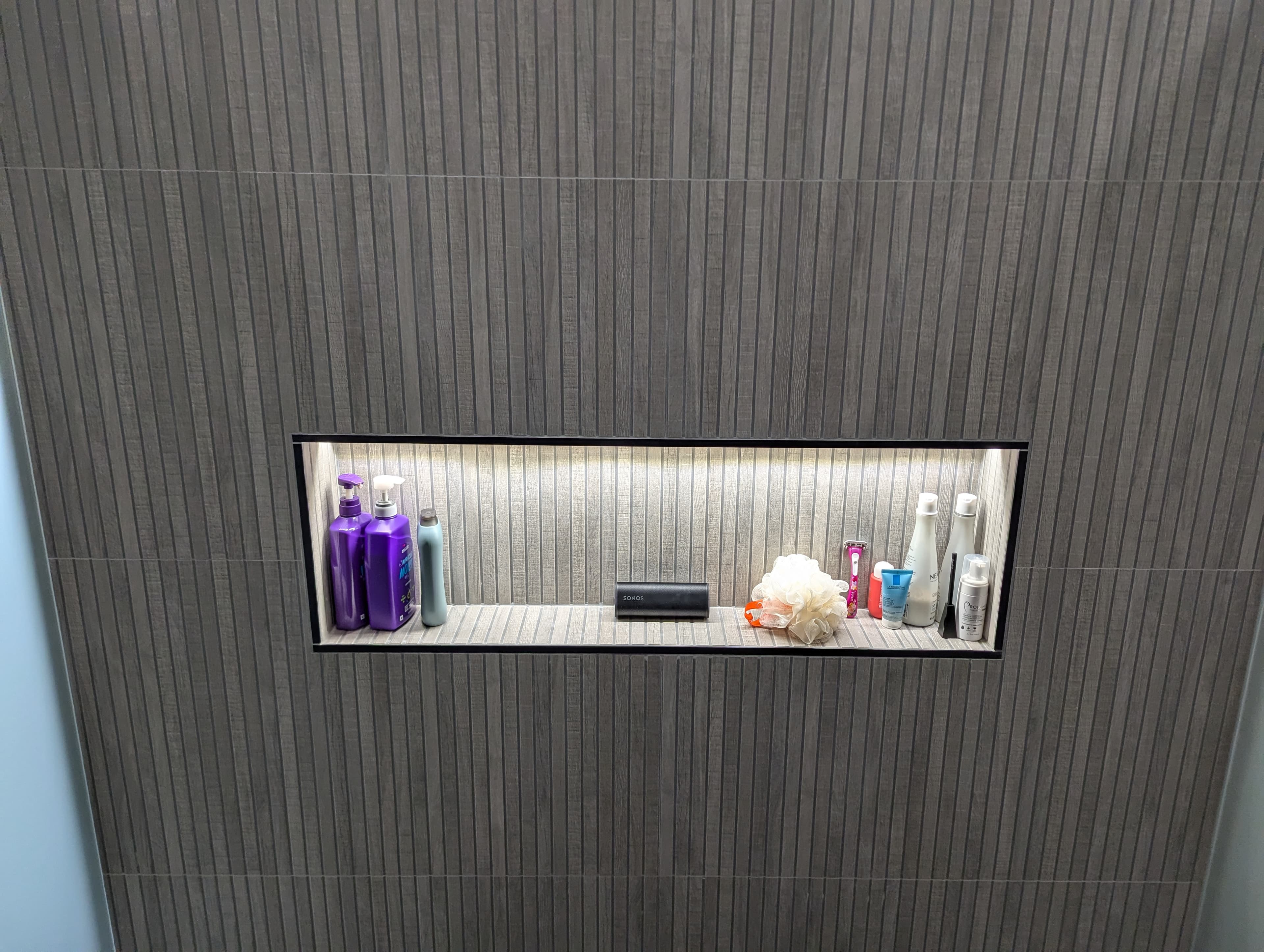 Sophisticated shower design showcasing vertical gray slat tile with a custom built-in shower niche featuring dramatic backlighting. The LED-illuminated niche provides both functional storage and striking visual appeal, creating a spa-like atmosphere in this modern bathroom renovation.