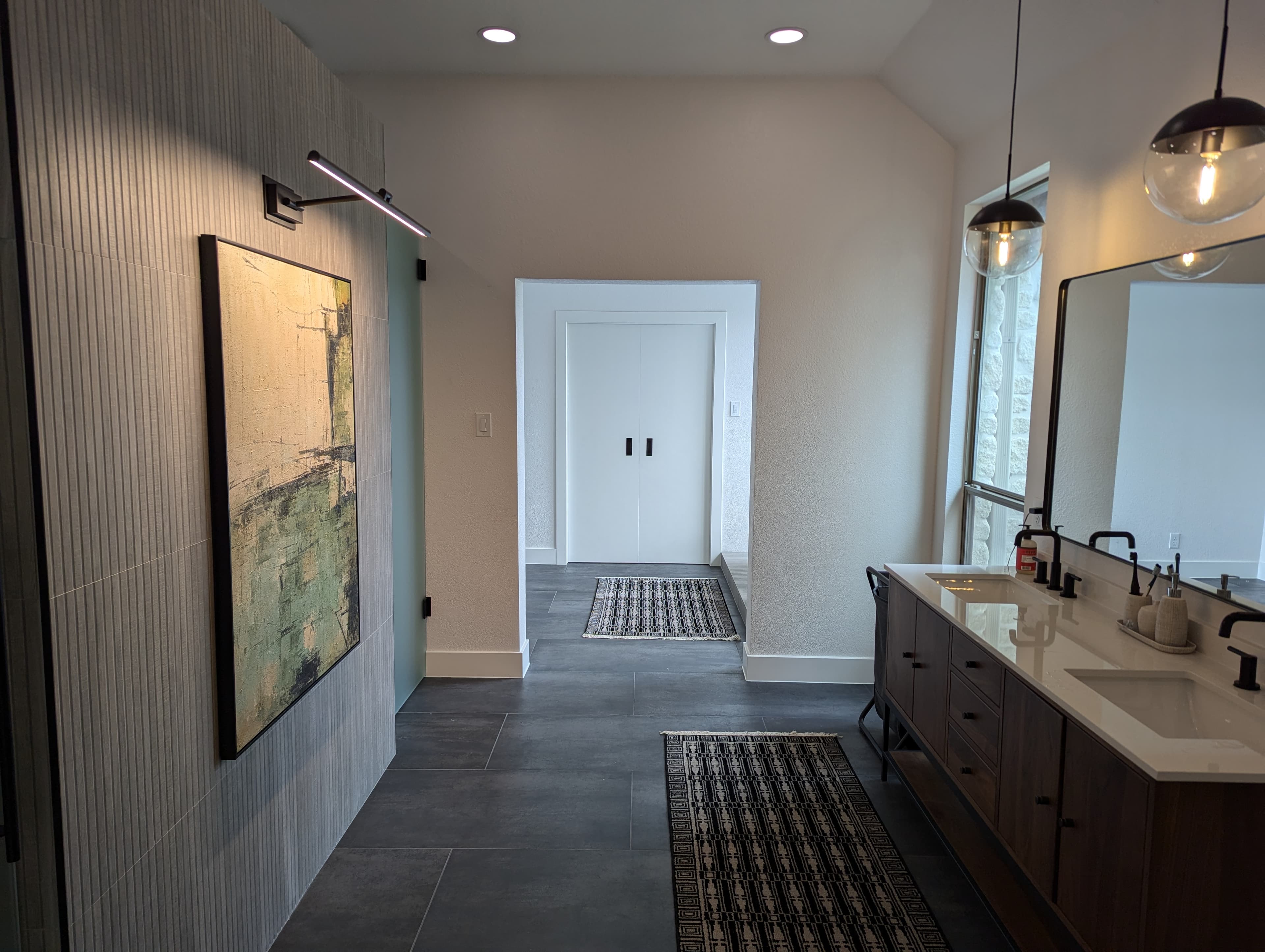 Elegant master bathroom renovation showcasing a dramatic vertical fluted tile accent wall, dark wood floating vanity with white quartz countertop, and high vaulted ceiling. The sophisticated design includes LED art lighting, modern globe pendant fixtures, and seamless flow to private areas, creating a spa-like retreat.