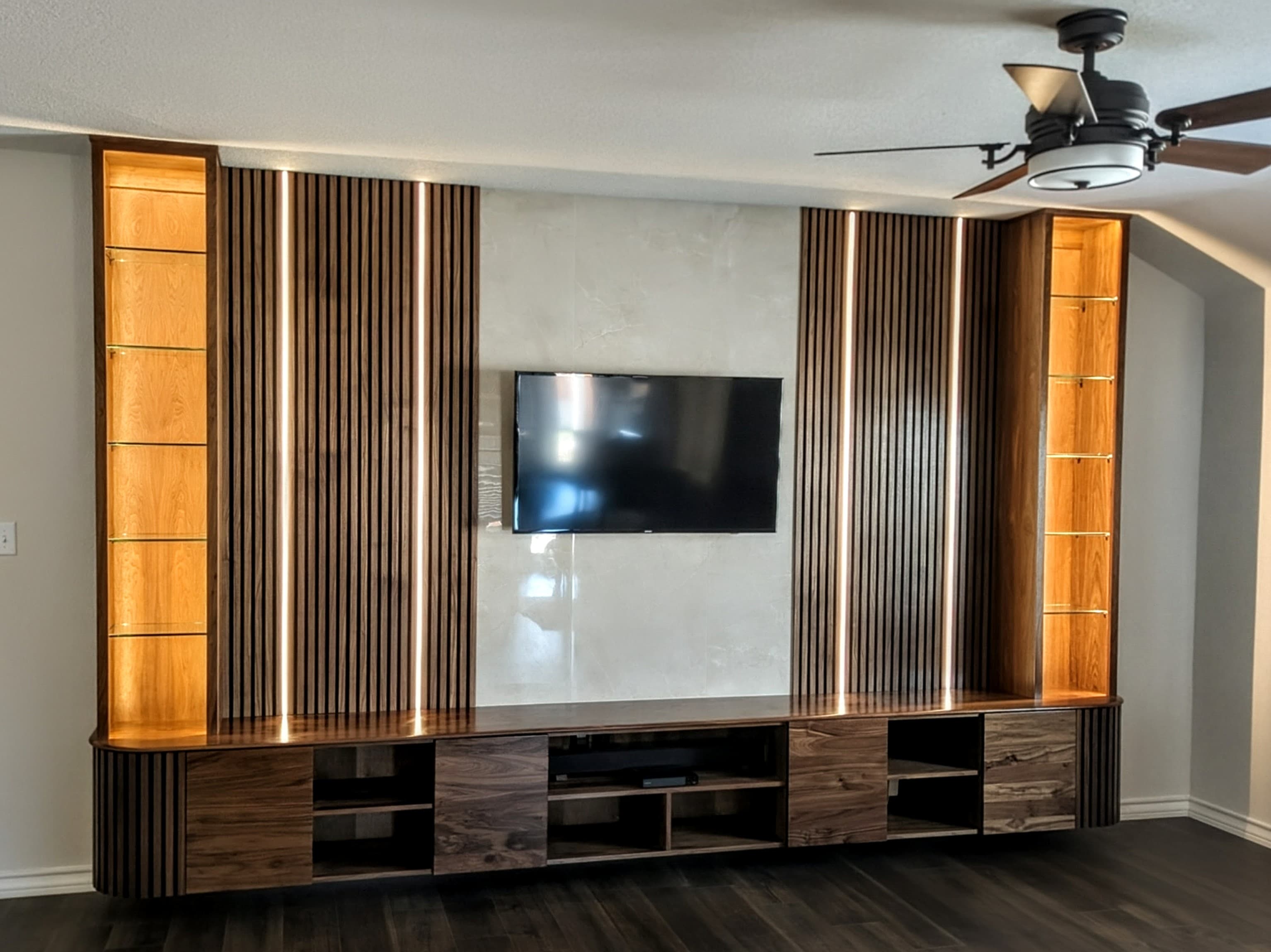 Show-stopping custom entertainment center featuring rich wood cabinetry with vertical slat wall panels and floating display shelves with integrated LED lighting. The modern design combines warm wood tones with clean lines, including lower cabinets with push-to-open drawers and cable management for a clutter-free media setup.