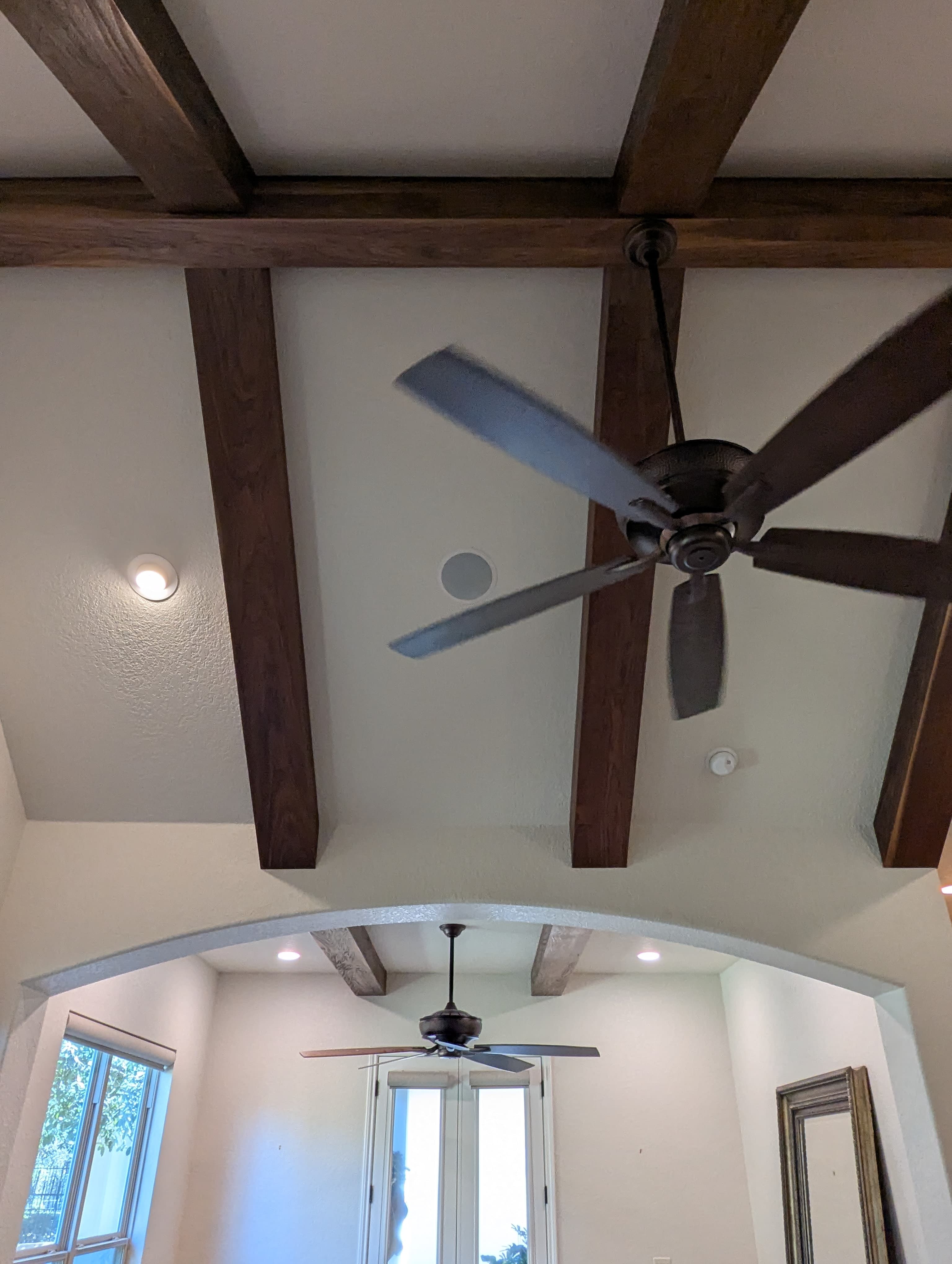 Custom walnut beam wraps installed over existing drywall beams in a vaulted ceiling near TPC Parkway in Stone Oak. The walnut was hand-distressed to achieve a rustic character and finished with a lacquer coating for durability. The wraps extend through an arched doorway, creating visual continuity between rooms.