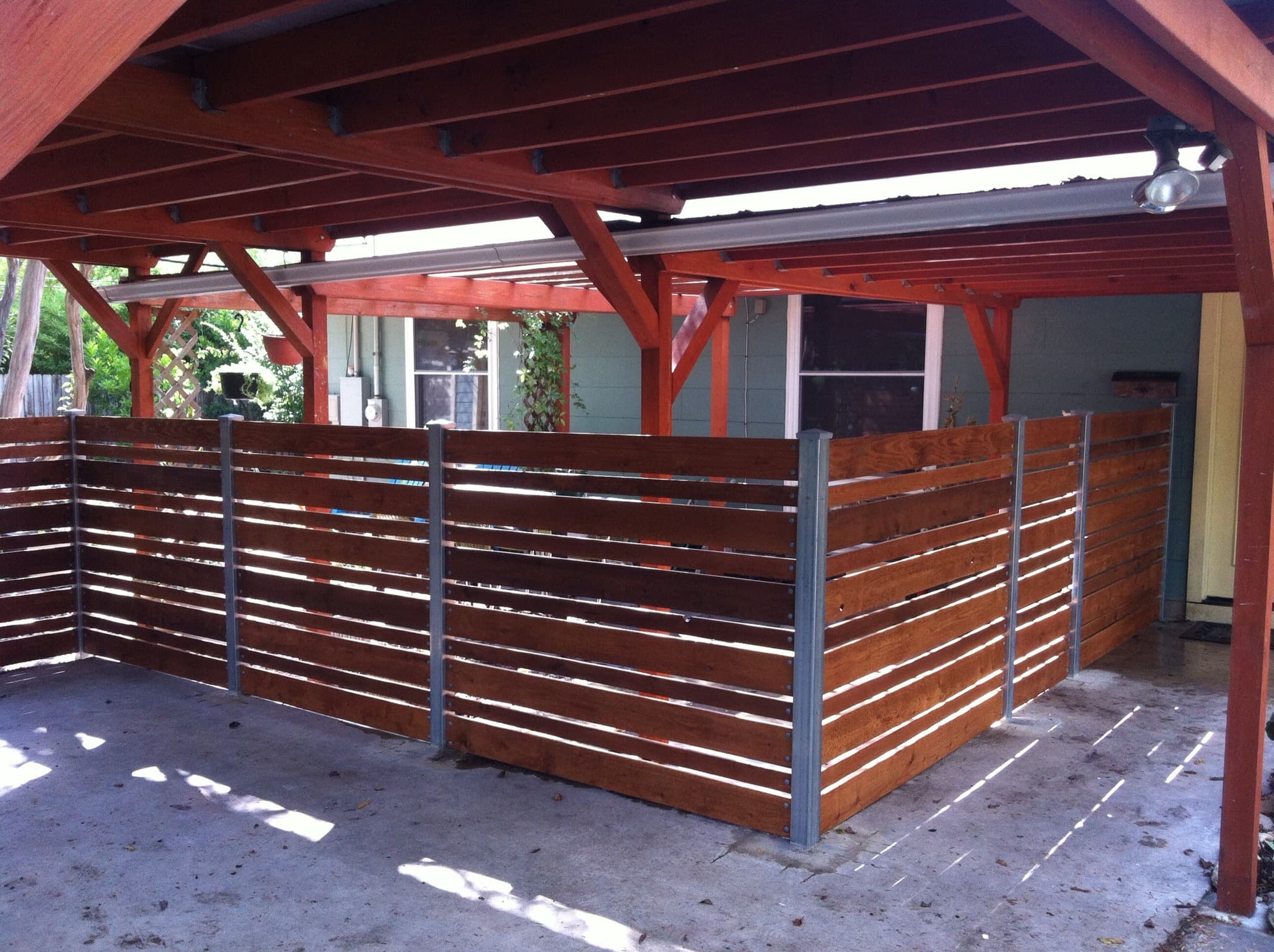 Dark-stained horizontal slat privacy screen set on galvanized steel posts, wrapping a carport to enclose the space without blocking airflow. The evenly spaced boards deliver a modern, architectural look that ties into the surrounding structure.